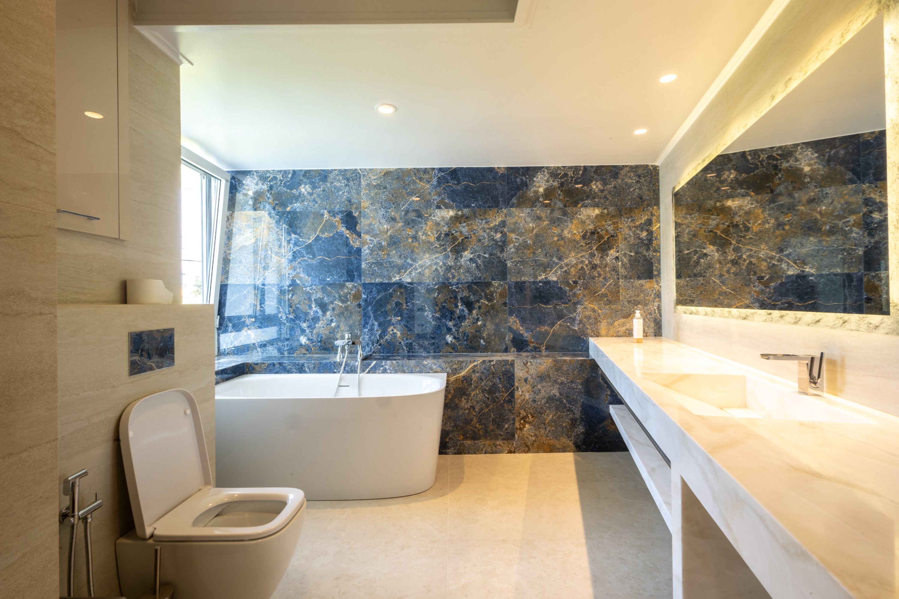 Freestanding bath against blue-black marble beside an illuminated vanity